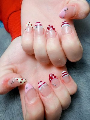 Medium nude almond nails with assorted red, pink, cream, and white French tips, polka dots, stripes, and silver studs.