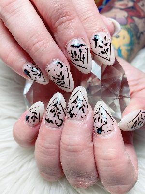Long nude almond nails carry detailed black botanical branches, bats, spiders, crescent moons and geometric frames.