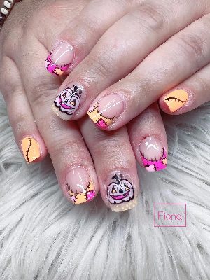 Short square nails with pink and peach stitched patchwork tips and smiling pale jack-o'-lantern accents.
