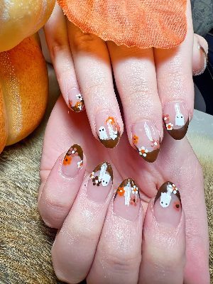 Glossy medium almond nails with a sheer pink base, brown tips, white ghosts, and small orange, white, and brown flowers.