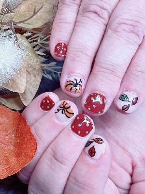 Short glossy round nails with red, pink, and orange designs featuring pumpkins, autumn leaves, and cream polka dots.
