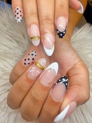 Long almond nails with nude-pink bases, black and white polka-dot French tips, gold studs and bands, a heart, and a painted letter R.