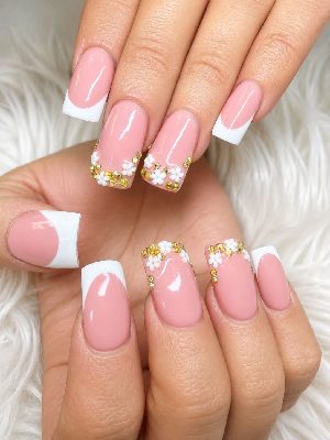 Long square pink nails with white French tips, raised white flower accents, and gold rhinestones on selected nails.