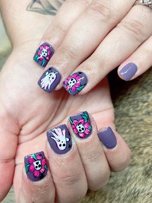 Short square purple nails with white ghosts, bright pink flowers containing skull faces, green leaves, and black starbursts.