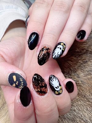 Medium almond nails in glossy black with white ghost characters, a skull face, bows, and copper glitter accents.