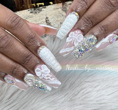 Extra-long coffin nails with nude-pink and white designs, sculpted bows, textured white panels, glitter edging, and oversized iridescent rhinestones.