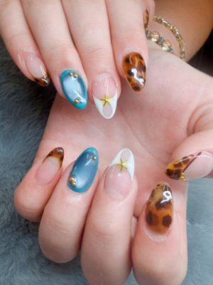 Almond nails with amber tortoise-shell tips, teal accent nails, white graphic accents, and small gold embellishments.